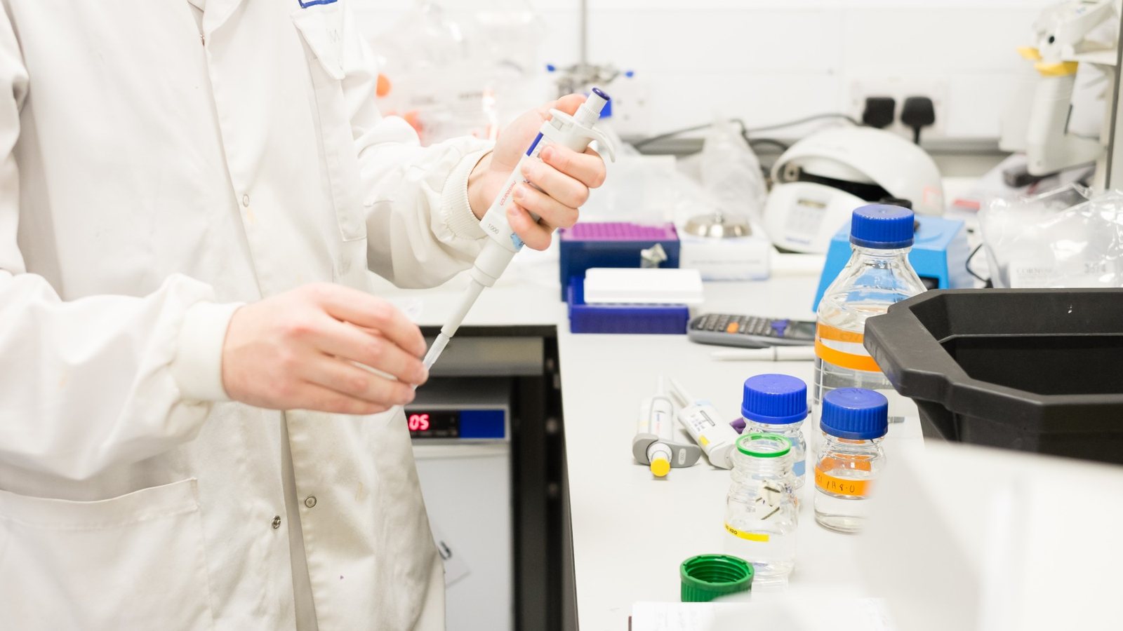 A scientist in a white lab coat using a pipette at a laboratory bench with sample bottles